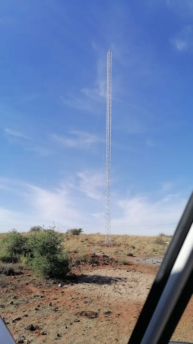A tall guyed lattice tower in dry, open scrubland under a blue sky.